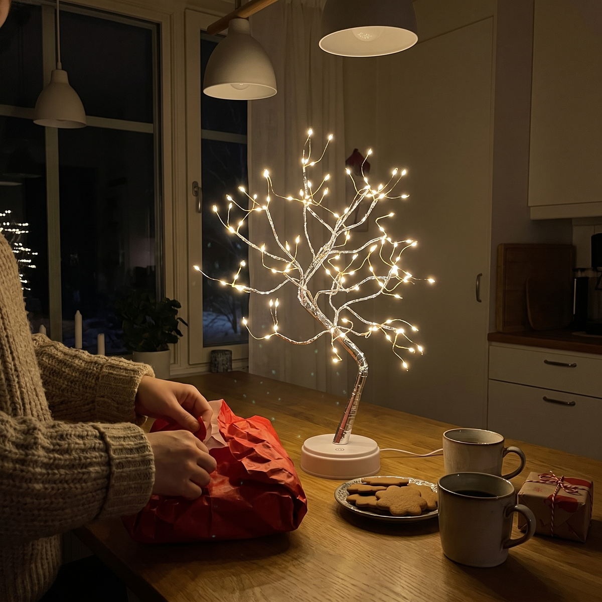 Händer öppnar en julklapp på köksbordet, Arbor lyser bredvid kaffemuggar och pepparkakor