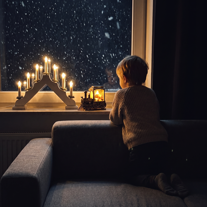 Ett barn på soffan tittar på Julius som lyser i fönstret bredvid adventsljusstaken en snöig kväll