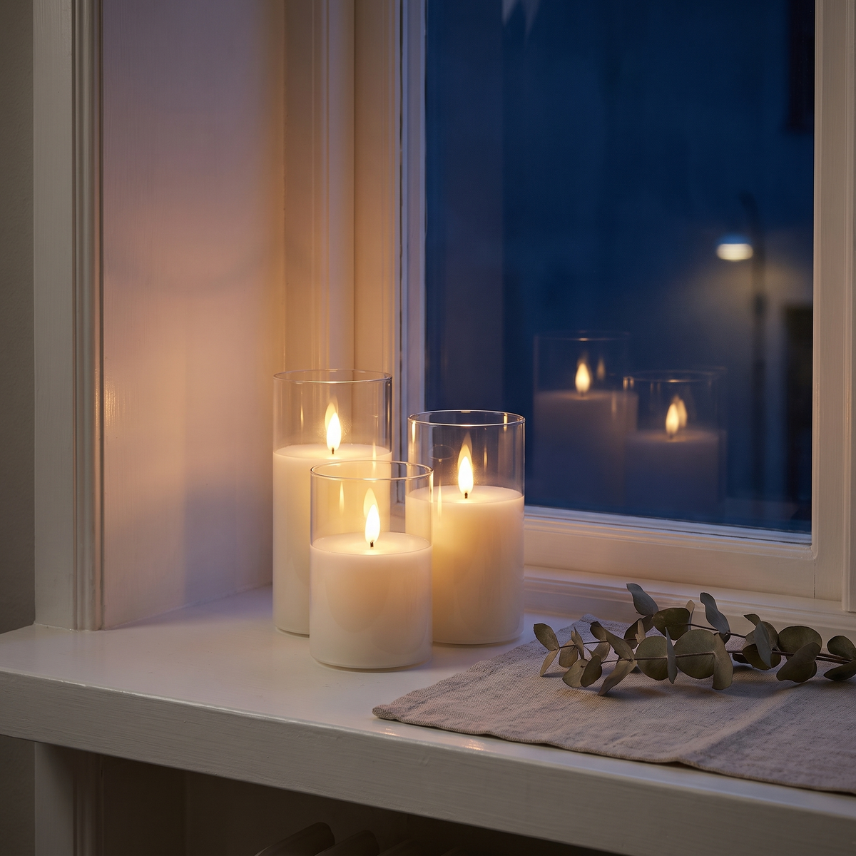 Cera i vitt: tre LED-blockljus i genomskinliga cylindrar tända i ett fönster en novemberkväll