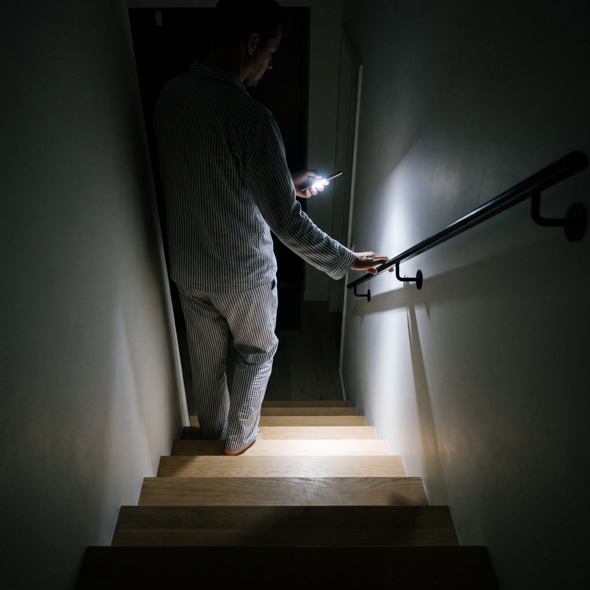 En person i pyjamas går nerför en mörk trappa med mobilens ficklampa och handen på ledstången